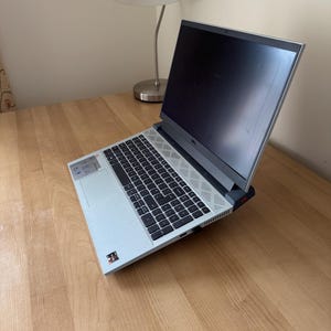May include: A light gray Dell laptop with a black keyboard and screen is open on a wooden desk. The laptop is angled towards the viewer, showcasing the keyboard and screen. A silver desk lamp is in the background.
