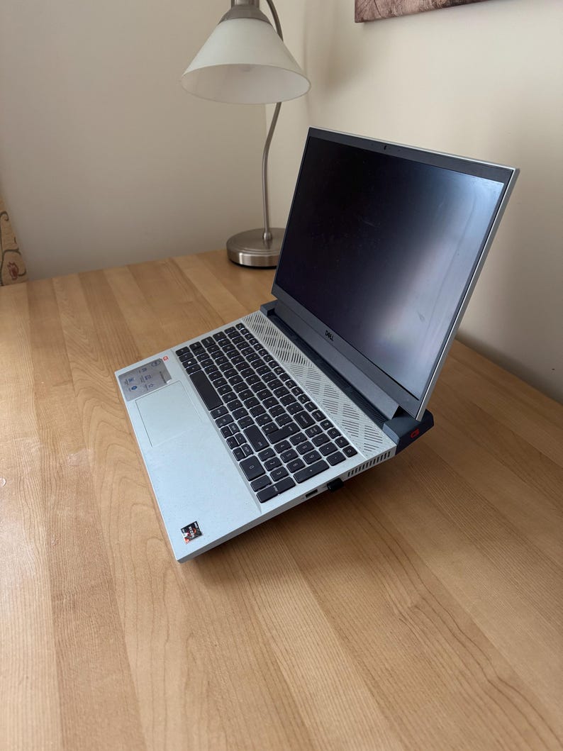 May include: A light gray Dell laptop with a black keyboard and screen is open on a wooden desk. The laptop is angled towards the viewer, showcasing the keyboard and screen. A silver desk lamp is in the background.