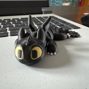 May include: A black 3D printed figurine of Toothless, the dragon from the How to Train Your Dragon movie series. The figurine has yellow eyes and is lying on its side with its wings spread out.