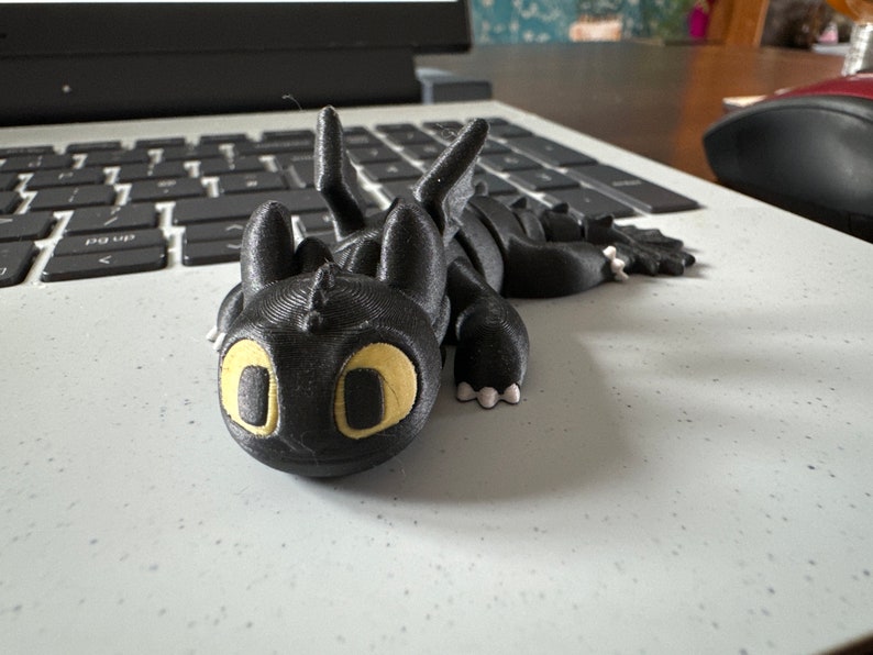 May include: A black 3D printed figurine of Toothless, the dragon from the How to Train Your Dragon movie series. The figurine has yellow eyes and is lying on its side with its wings spread out.