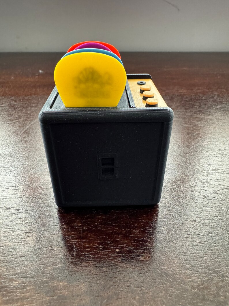 May include: A black guitar amplifier with a built-in pick holder. The pick holder has four slots and is filled with colorful guitar picks.