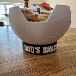 Op de afbeelding: Een witte, voetbalvormige snackkom met een zwarte basis. De basis leest "DAD'S SNACKS" in witte letters. De kom is gevuld met diverse snackverpakkingen.
