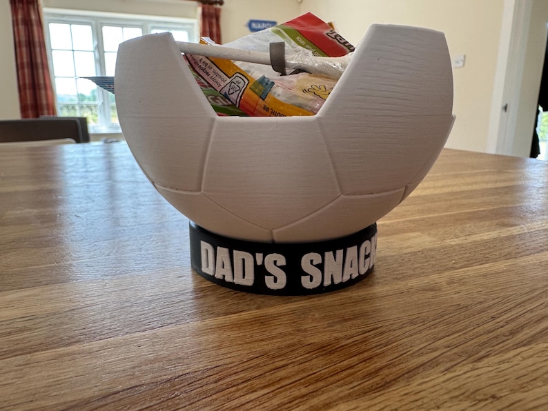 Op de afbeelding: Een witte, voetbalvormige snackkom met een zwarte basis. De basis leest "DAD'S SNACKS" in witte letters. De kom is gevuld met diverse snackverpakkingen.