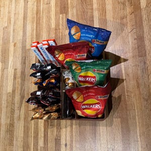 May include: A brown tray filled with various snacks, including bags of Walkers crisps in red, green, and blue packaging, and chocolate bars. The snacks are arranged on a wooden surface.