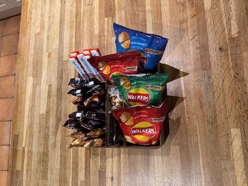 May include: A brown tray filled with various snacks, including bags of Walkers crisps in red, green, and blue packaging, and chocolate bars. The snacks are arranged on a wooden surface.