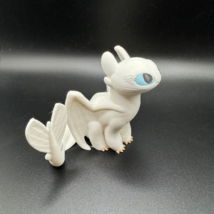 May include: White 3D printed figurine of a dragon with blue eyes and a curled tail. The dragon is standing on a black surface.