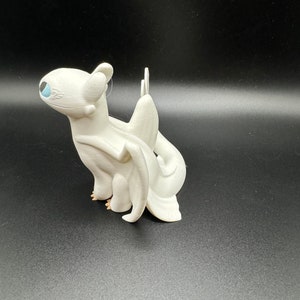 May include: White figurine of a dragon with a blue eye. The dragon has wings and a tail. The figurine is made of plastic.