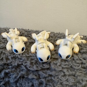 May include: Three white Light Fury dragons from the How to Train Your Dragon movie series, lying on a gray fuzzy surface. The dragons have blue eyes and are facing the camera.
