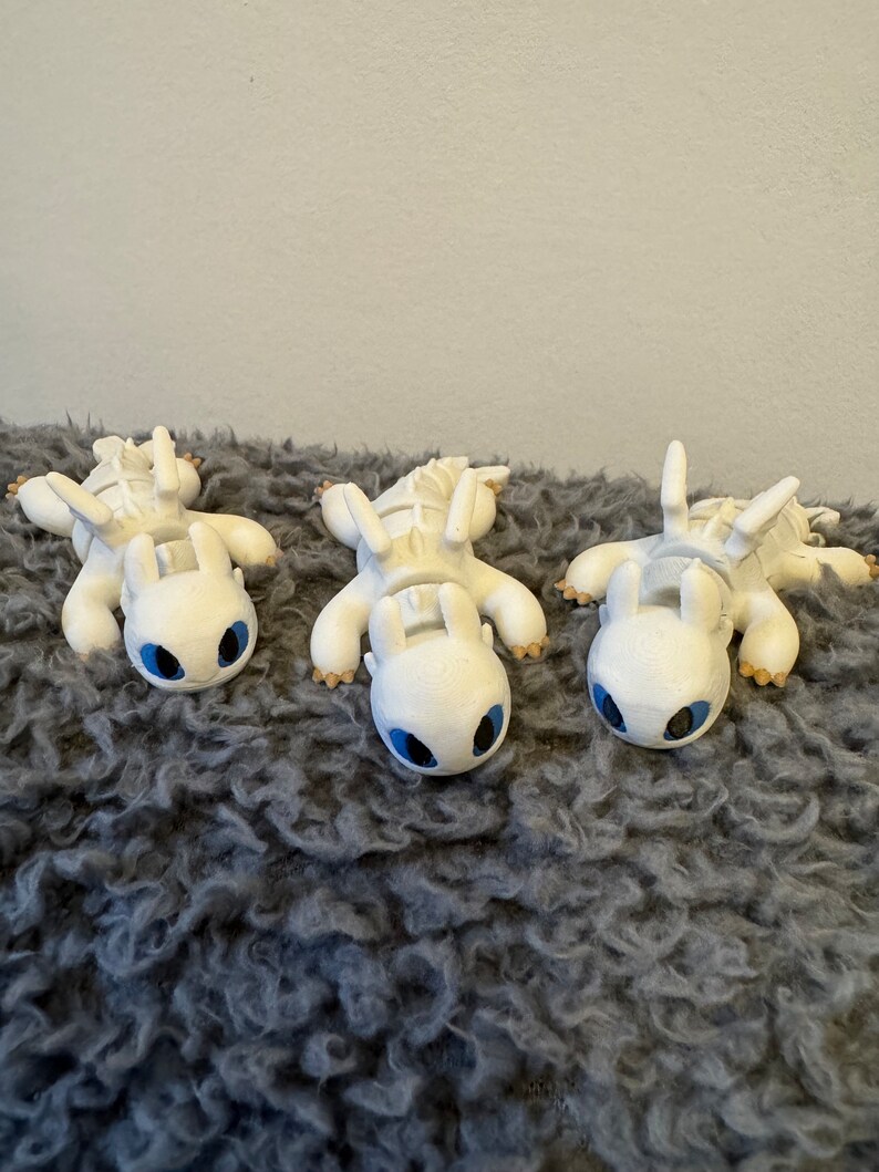 May include: Three white Light Fury dragons from the How to Train Your Dragon movie series, lying on a gray fuzzy surface. The dragons have blue eyes and are facing the camera.