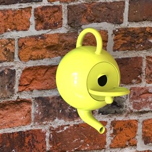 May include: A bright yellow birdhouse with a round body, a small spout, and a handle. The birdhouse is mounted on a weathered brick wall. The birdhouse has a small perch and a circular opening for the birds.