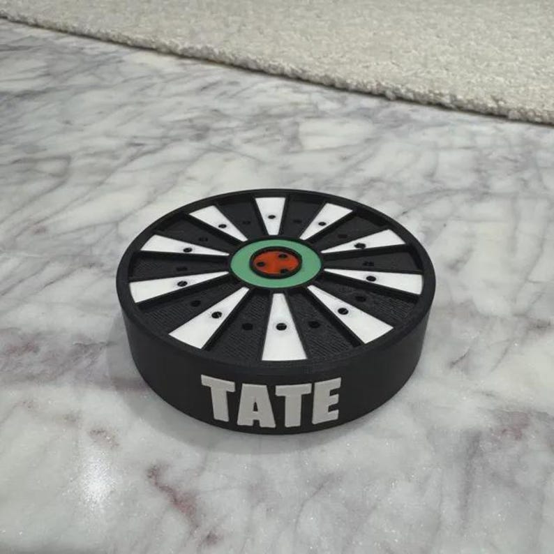 May include: A black and white dartboard-style object with the word "TATE" in white block letters on the side. The center has a green ring with a red and orange center. The object is circular.