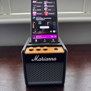 May include: A black and gray portable speaker with a yellow knob and the word "Marianna" printed on the front. A smartphone is sitting on top of the speaker and is playing a podcast.