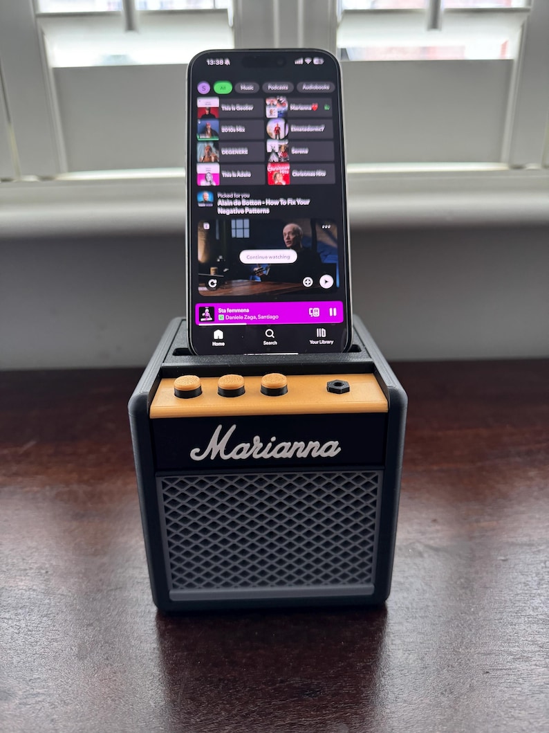 May include: A black and gray portable speaker with a yellow knob and the word "Marianna" printed on the front. A smartphone is sitting on top of the speaker and is playing a podcast.