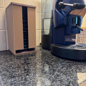 May include: A brown, vertical coffee pod holder with a ribbed design, next to a blue espresso machine. The holder is rectangular and appears to be made of plastic. The espresso machine has a water reservoir and a spout.