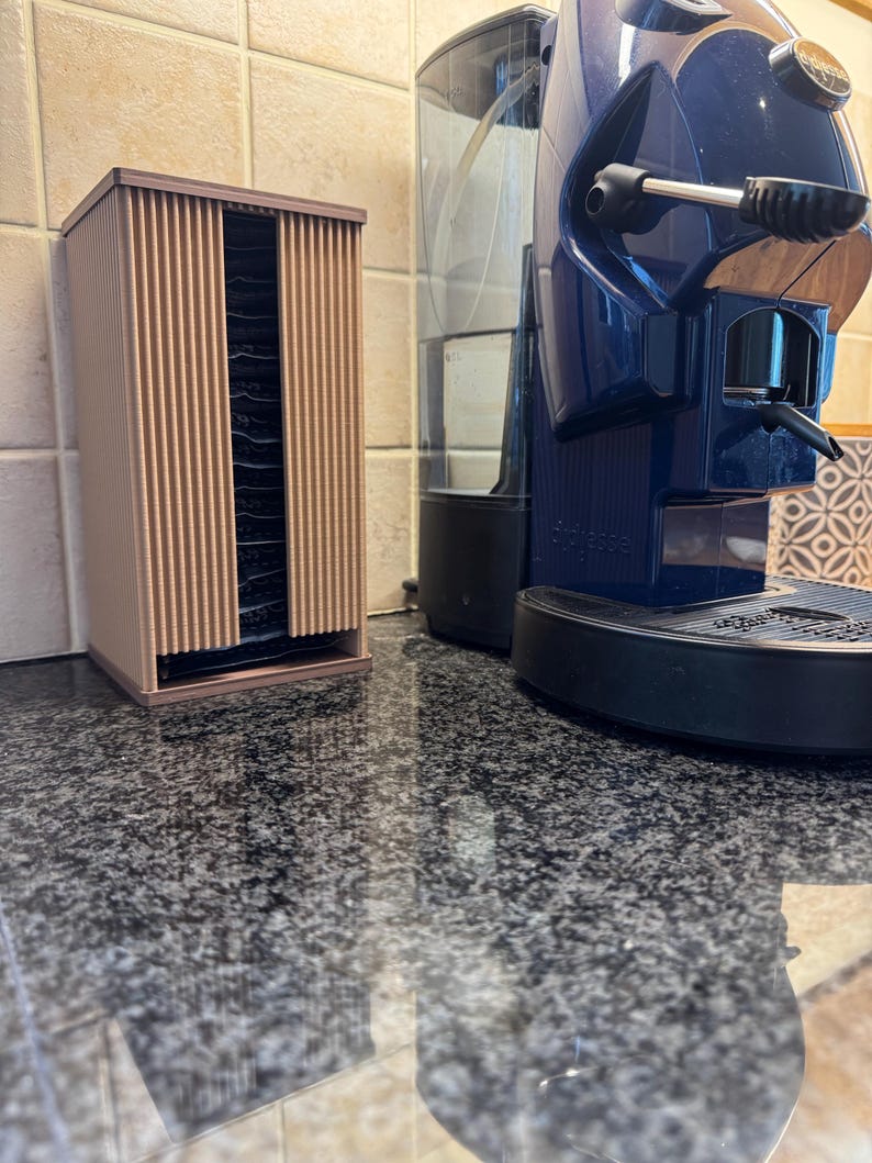 May include: A brown, vertical coffee pod holder with a ribbed design, next to a blue espresso machine. The holder is rectangular and appears to be made of plastic. The espresso machine has a water reservoir and a spout.