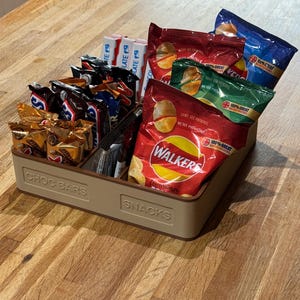 May include: A beige rectangular snack organizer filled with various individually wrapped snacks. The organizer is divided into sections labeled "CHOC BARS" and "SNACKS". The snacks include chocolate bars and bags of chips.