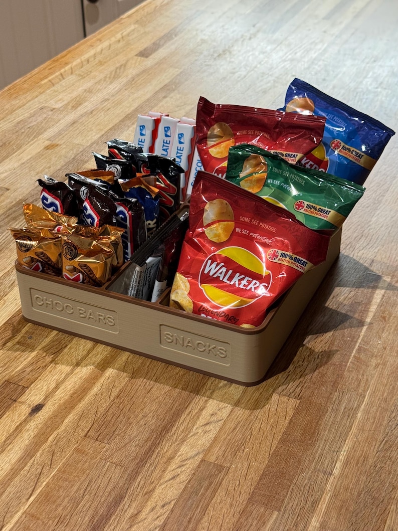 May include: A beige rectangular snack organizer filled with various individually wrapped snacks. The organizer is divided into sections labeled "CHOC BARS" and "SNACKS". The snacks include chocolate bars and bags of chips.