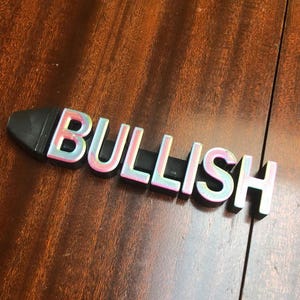 May include: A black and iridescent plastic word block spelling "BULLISH" on a wooden surface.