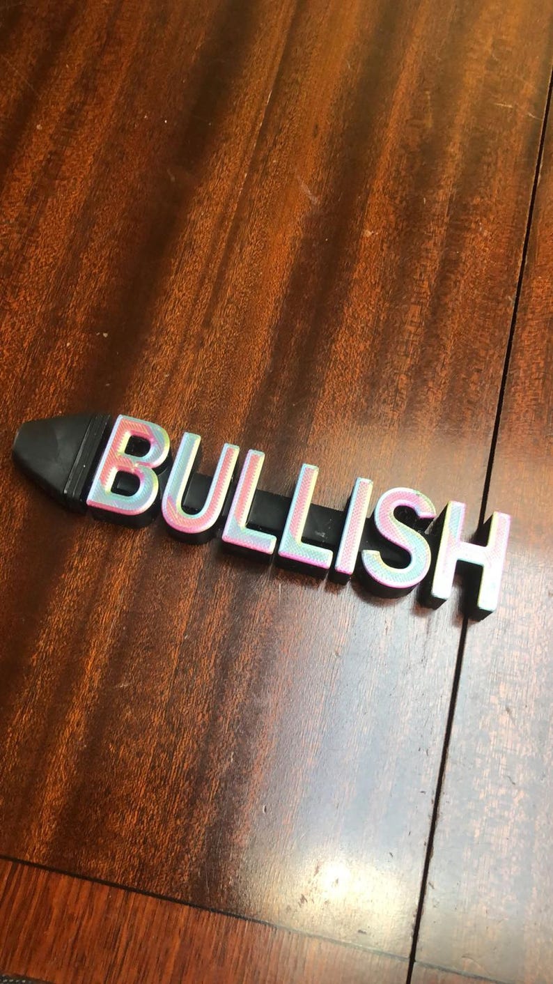 May include: A black and iridescent plastic word block spelling "BULLISH" on a wooden surface.