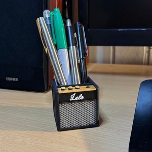 May include: A black and gold miniature amplifier-shaped desk organizer with the word "Lele" on the front. It holds a variety of pens and markers.