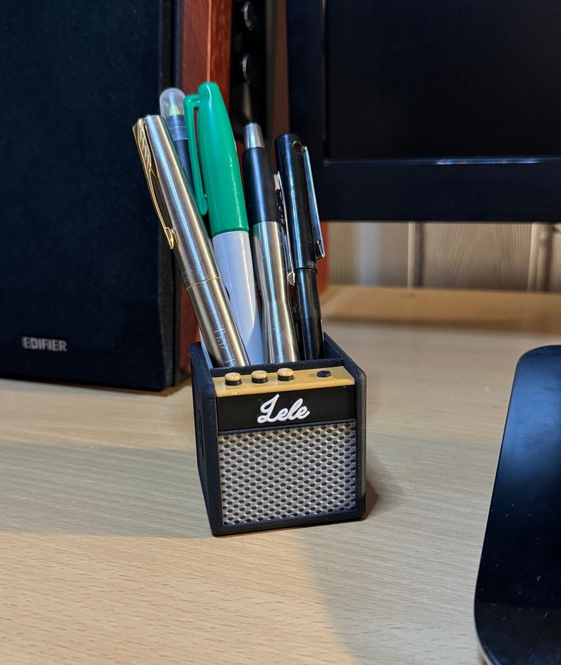 May include: A black and gold miniature amplifier-shaped desk organizer with the word "Lele" on the front. It holds a variety of pens and markers.