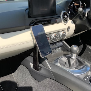 K&ouml;nnte beinhalten: Innenansicht eines Autos mit einem Smartphone, das an einer schwarzen Halterung befestigt ist. Das Telefon befindet sich in der N&auml;he des Schalthebels, mit einem schwarzen Infotainment-Bildschirm dar&uuml;ber. Das Armaturenbrett ist cremefarben mit schwarzen Akzenten.