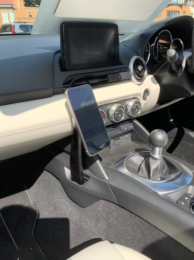 K&ouml;nnte beinhalten: Innenansicht eines Autos mit einem Smartphone, das an einer schwarzen Halterung befestigt ist. Das Telefon befindet sich in der N&auml;he des Schalthebels, mit einem schwarzen Infotainment-Bildschirm dar&uuml;ber. Das Armaturenbrett ist cremefarben mit schwarzen Akzenten.