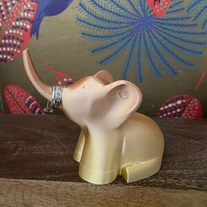 Op de afbeelding: Een gouden olifantenfiguur met een ringhouder op zijn slurf. De olifant staat op een houten oppervlak.