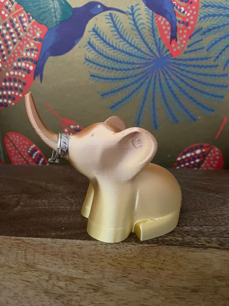 Op de afbeelding: Een gouden olifantenfiguur met een ringhouder op zijn slurf. De olifant staat op een houten oppervlak.
