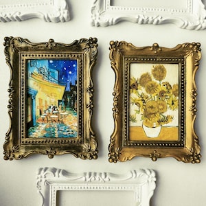Può includere: Due cornici rettangolari dorate e decorate contenenti stampe artistiche in miniatura. La stampa a sinistra raffigura una scena di un caffè notturno nei toni del blu e del giallo. Quella a destra mostra un bouquet di girasoli in un vaso bianco.