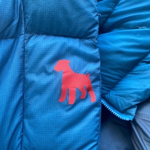 May include: Close-up of a bright blue puffer jacket with a red dog silhouette on the front. The jacket has a quilted texture and a zipper closure. The dog graphic is a solid, simple design.