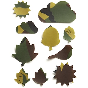 Puede incluir: Una colección de diez formas planas troqueladas con patrones de camuflaje en tonos verde oliva, marrón, negro y amarillo. Las formas incluyen un sol, nubes, hojas, una bellota y un pájaro, todos con una superficie lisa y mate.