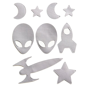 May include: A collection of nine silver-colored fabric patches featuring space-themed shapes including two crescent moons, three five-pointed stars, two alien heads with large oval eyes, one upright rocket ship, and one horizontal rocket ship design.