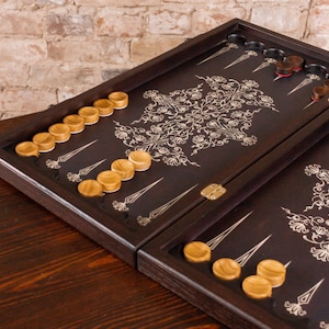 Könnte beinhalten: Ein dunkelbraunes Backgammon-Brett mit einem dekorativen floralen Design in einer helleren Farbe. Das Brett ist geöffnet und zeigt Spielsteine aus Holz in hellen und dunklen Tönen. Die Spielsteine sind auf dem Brett angeordnet und spielbereit.
