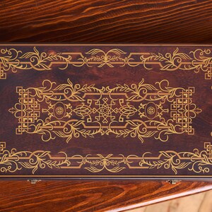 Könnte beinhalten: Eine rechteckige Holzkiste mit dunkelbraunem Finish und aufwendigen goldfarbenen floralen und geometrischen Designs. Die Kiste ist geschlossen und scheint ein dekorativer Gegenstand oder ein Aufbewahrungsbehälter zu sein. Das Design ist symmetrisch und verziert.