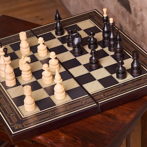 Könnte beinhalten: Ein Schachspiel mit schwarzen und cremefarbenen Figuren auf einem dunkelbraunen und beigefarbenen Schachbrett. Das Brett steht auf einem passenden Holztisch. Der Rand des Bretts hat ein dekoratives Golddesign.