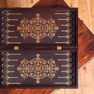 Könnte beinhalten: Geöffnetes Backgammon-Spielbrett mit dunkelbraunem Finish und goldenen Akzenten. Das Spielbrett hat ein florales Design in der Mitte und an den Rändern. Das Spielbrett wird auf einem Holztisch präsentiert.