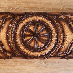 May include: A rectangular wooden backgammon board with detailed carved floral and geometric patterns. The board has a dark brown border and a lighter, warm brown interior, creating a contrast. The carvings add a touch of elegance to the board.