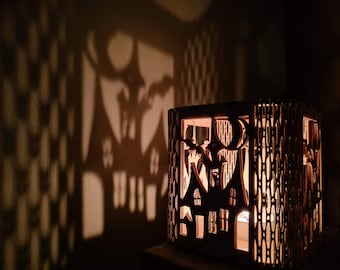 May include: A brown, laser-cut candle holder featuring a cityscape design. The candle holder projects shadows of buildings, a moon, and bats onto the wall. The warm light from the candle illuminates the intricate cutouts, creating a cosy ambience.