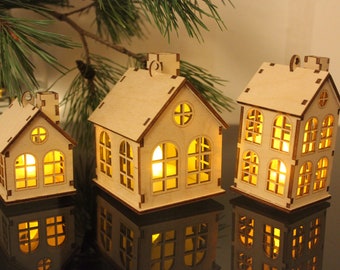 Puede incluir: Tres portavelas de madera con forma de casa con luces interiores brillantes. Las casas tienen múltiples ventanas y chimeneas. Los portavelas están hechos de madera de color claro y se asientan sobre una superficie reflectante. Una rama de pino verde está en el fondo.