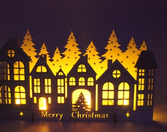 Puede incluir: Una escena de pueblo navideño de madera con casas, árboles y la inscripción "Merry Christmas" iluminados. Las casas tienen ventanas y puertas recortadas, con un cálido resplandor amarillo. Los árboles se recortan contra la luz.