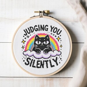 Könnte beinhalten: Kreuzstich-Stickrahmen mit einem schwarzen Kätzchen auf einer Wolke mit Regenbogen und Sternen. Der Text lautet "JUDGING YOU SILENTLY" in schwarzen Buchstaben. Das Kunstwerk befindet sich in einem Holzstickrahmen.