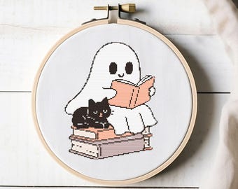 May include: A cross-stitch embroidery hoop featuring a cartoon ghost reading a book, with a black cat sitting on a stack of books. The design is in shades of white, black, and peach, set against a white background.