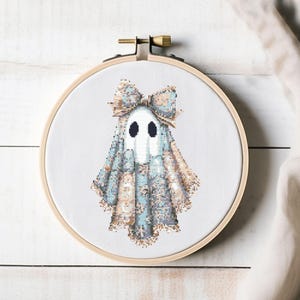May include: A cross-stitch embroidery of a ghost wearing a bow, set in a wooden hoop. The ghost is white with black eyes and a blue and tan patterned outfit. The bow is tan and blue. The background is white.