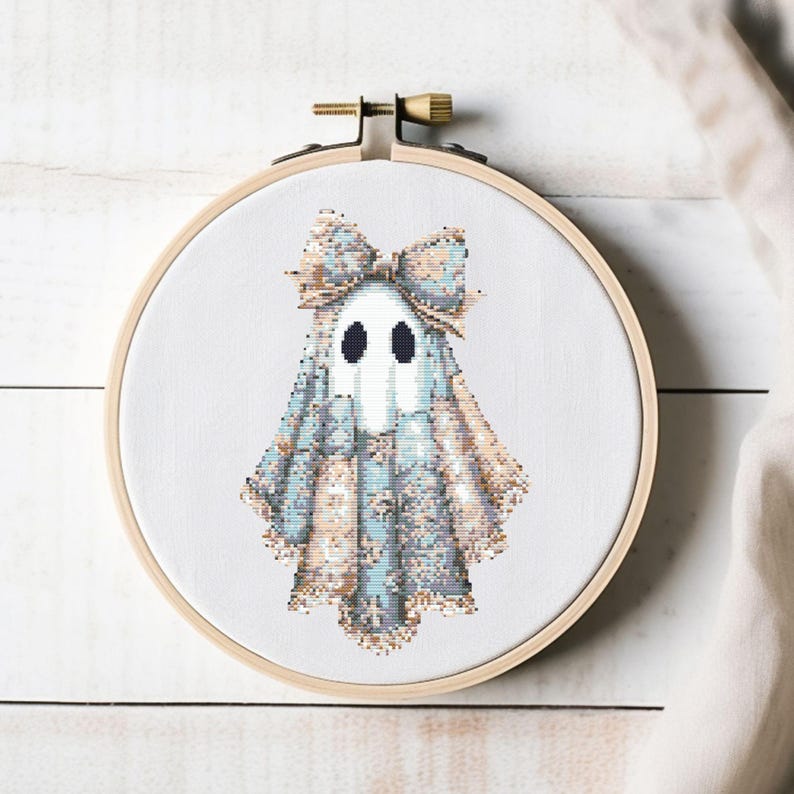 May include: A cross-stitch embroidery of a ghost wearing a bow, set in a wooden hoop. The ghost is white with black eyes and a blue and tan patterned outfit. The bow is tan and blue. The background is white.