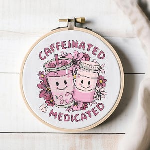 Könnte beinhalten: Ein Kreuzstich-Stickrahmen mit den Worten "Caffeinated Medicated" in Rosa. Das Design zeigt zwei lächelnde rosa Trinkbecher, umgeben von rosa Blumen. Der Rahmen ist aus Holz.