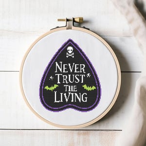Könnte beinhalten: Kreuzstich-Stickerei in einem Holzreifen. Das Design zeigt eine schwarze und lila Planchette mit dem Text "NEVER TRUST THE LIVING", einen Totenkopf und Fledermäuse. Die Stickerei befindet sich auf weißem Stoff.