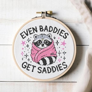 Könnte beinhalten: Ein Stickrahmen mit einem Waschbären, der in eine rosa Decke gehüllt ist. Der Text "EVEN BADDIES GET SADDIES" ist über und unter dem Waschbären gestickt. Rosa Sterne und Punkte umgeben den Waschbären, alles in einem Holzrahmen.