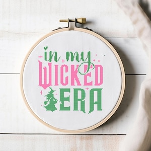 Könnte beinhalten: Ein Kreuzstich-Stickrahmen mit dem Text "in my WICKED ERA". Der Text ist in Rosa und Grün gehalten, mit einer Hexen-Grafik. Der Rahmen ist aus Holz und hat einen Messingverschluss.