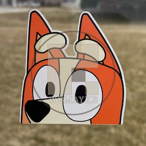 May include: A die-cut vinyl sticker featuring a cartoon dog character with orange fur, cream-colored accents, and large white eyes with black pupils. The character has pointed ears and a black nose. The text WEMAYKE is visible in the center.